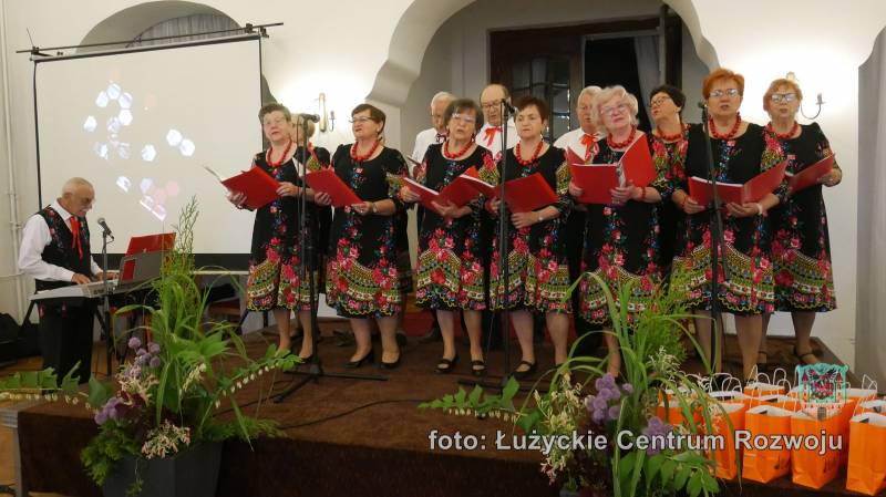 Grupa kobiet i mężczyzn w strojach stylizowanych na ludowe stoi śpiewa na scenie